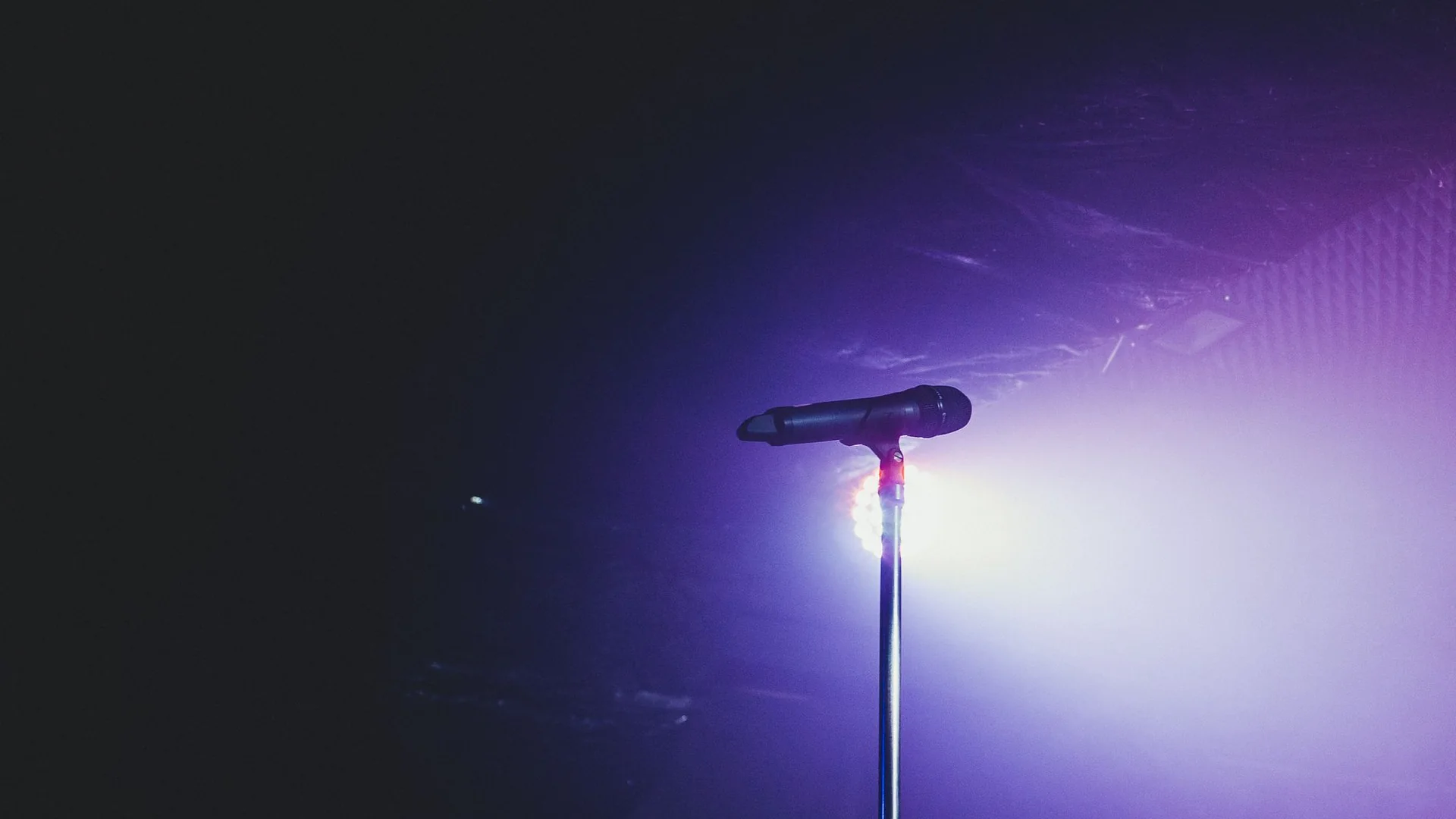 A microphone on a stand lit by a purple stage spotlight against a dark theatre ceiling