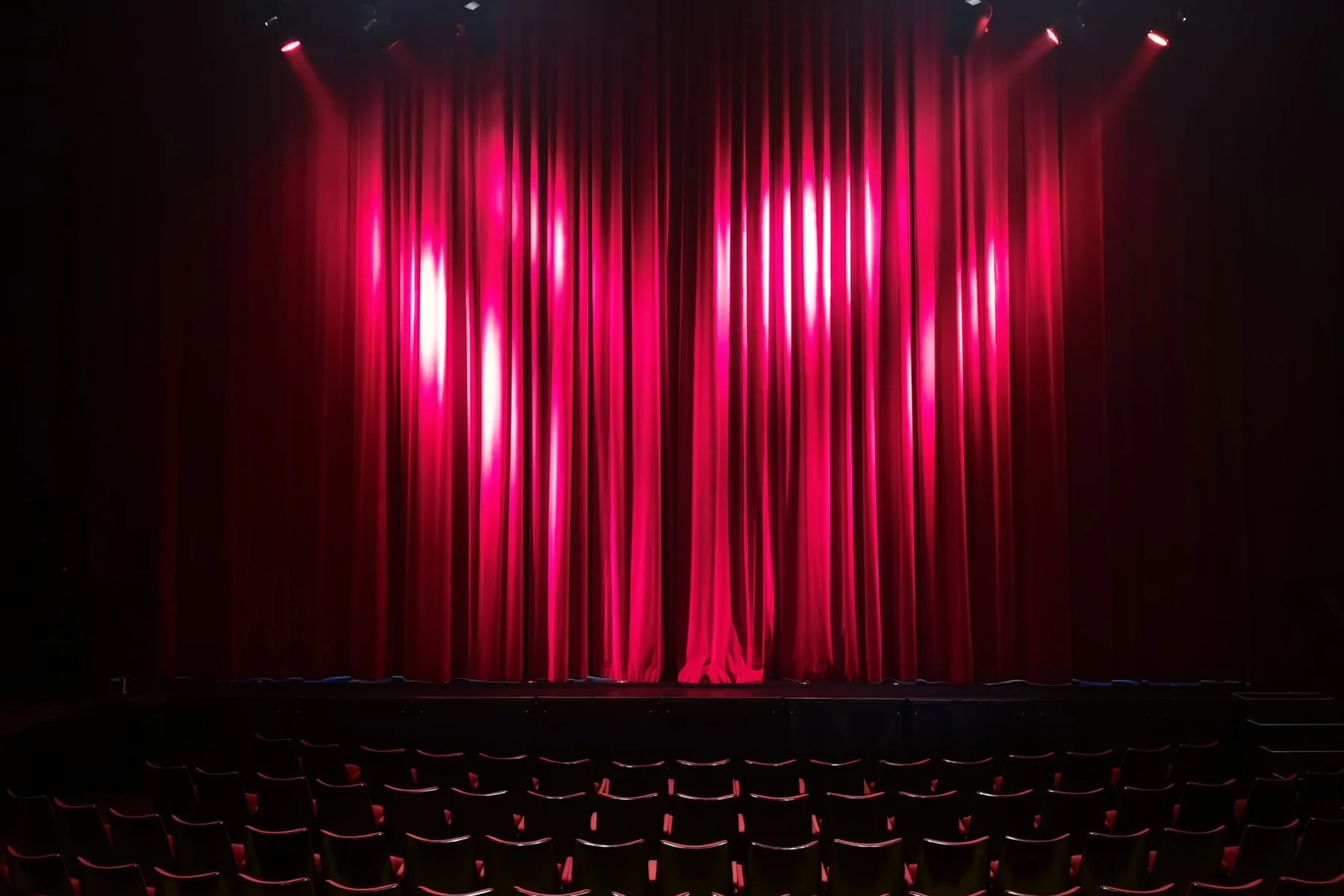Empty theatre seats facing a red stage curtain lit by spotlights